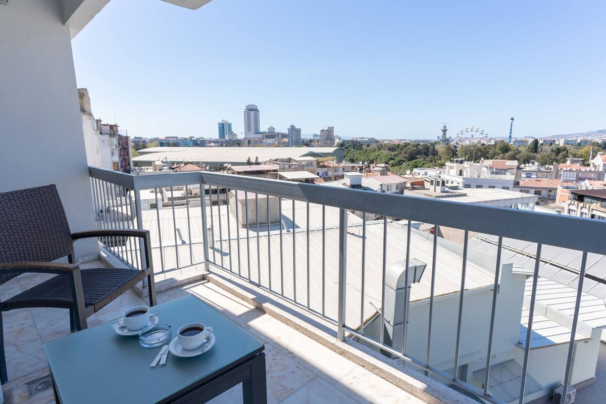 Un balcon avec une table et des chaises, donnant sur un paysage urbain avec des bâtiments et un ciel dégagé.