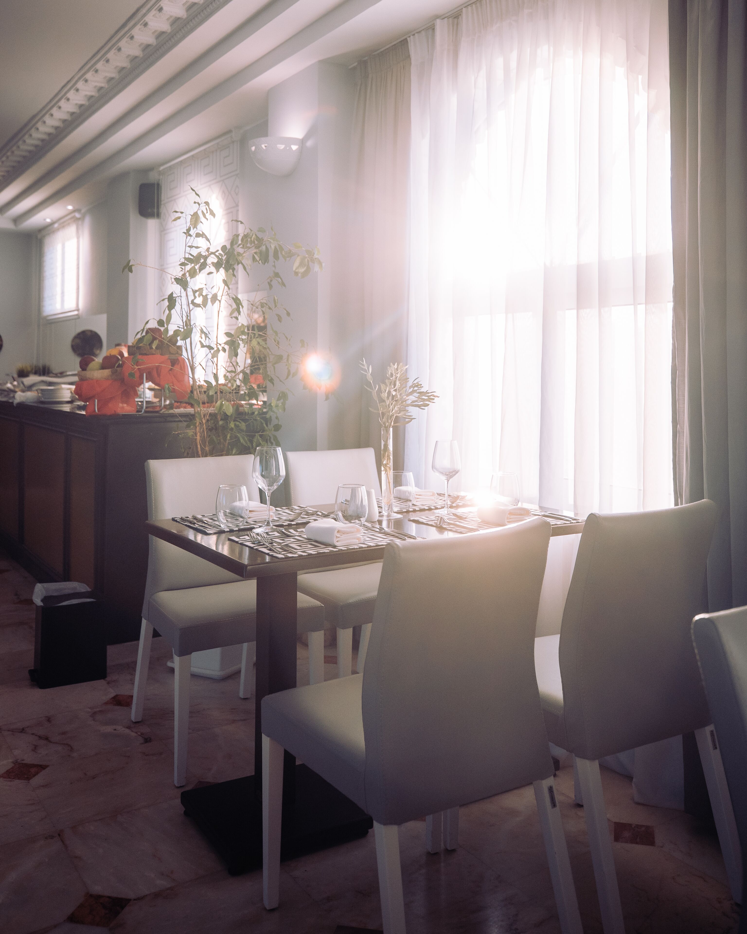 A dining area with a table set for four, chairs, a potted plant, and a window with sheer curtains.