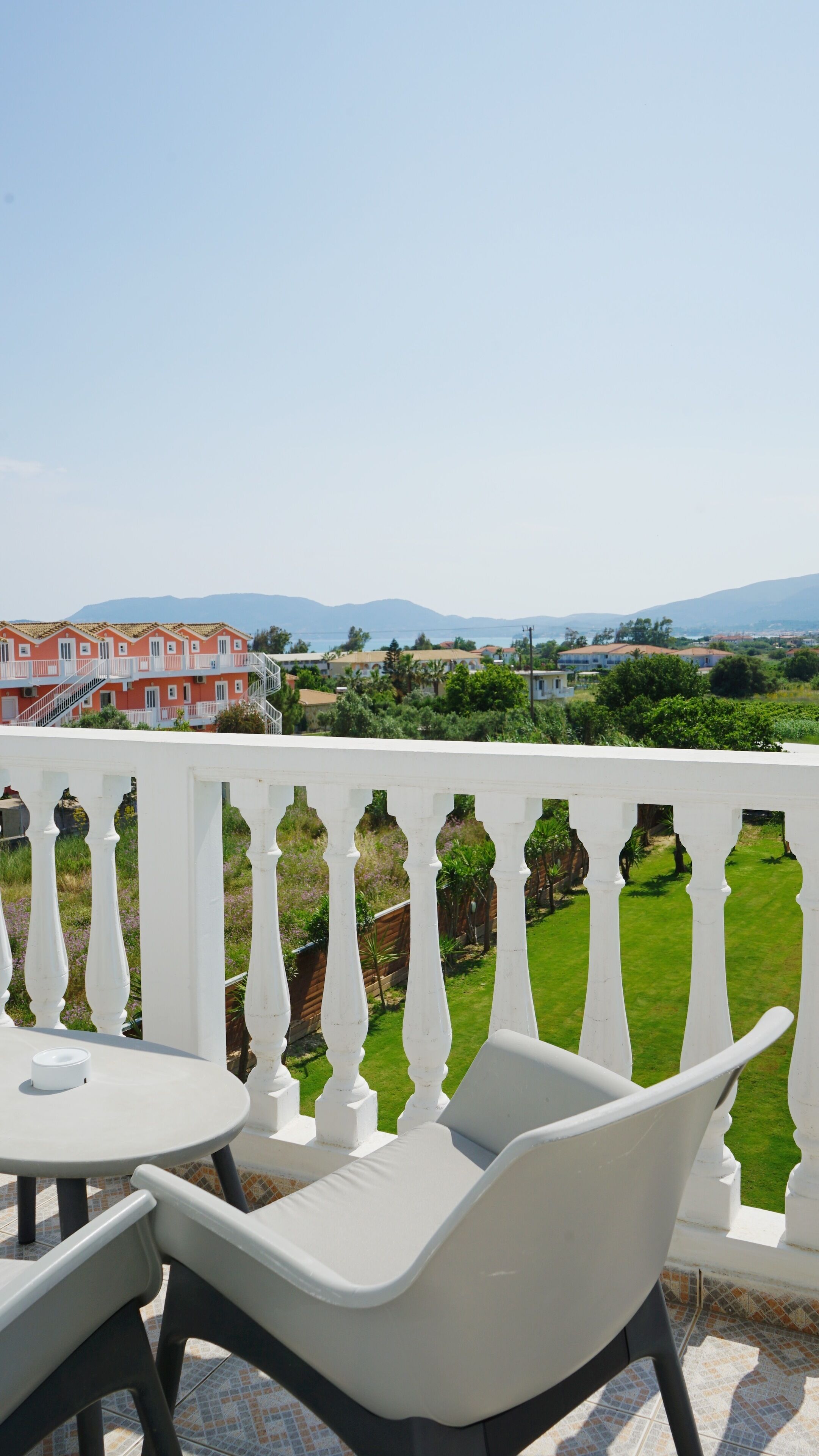 A balcony with white balustrades, a round table, and two white chairs. In the background, there are buildings with red and white facades, and a view of green fields and mountains.