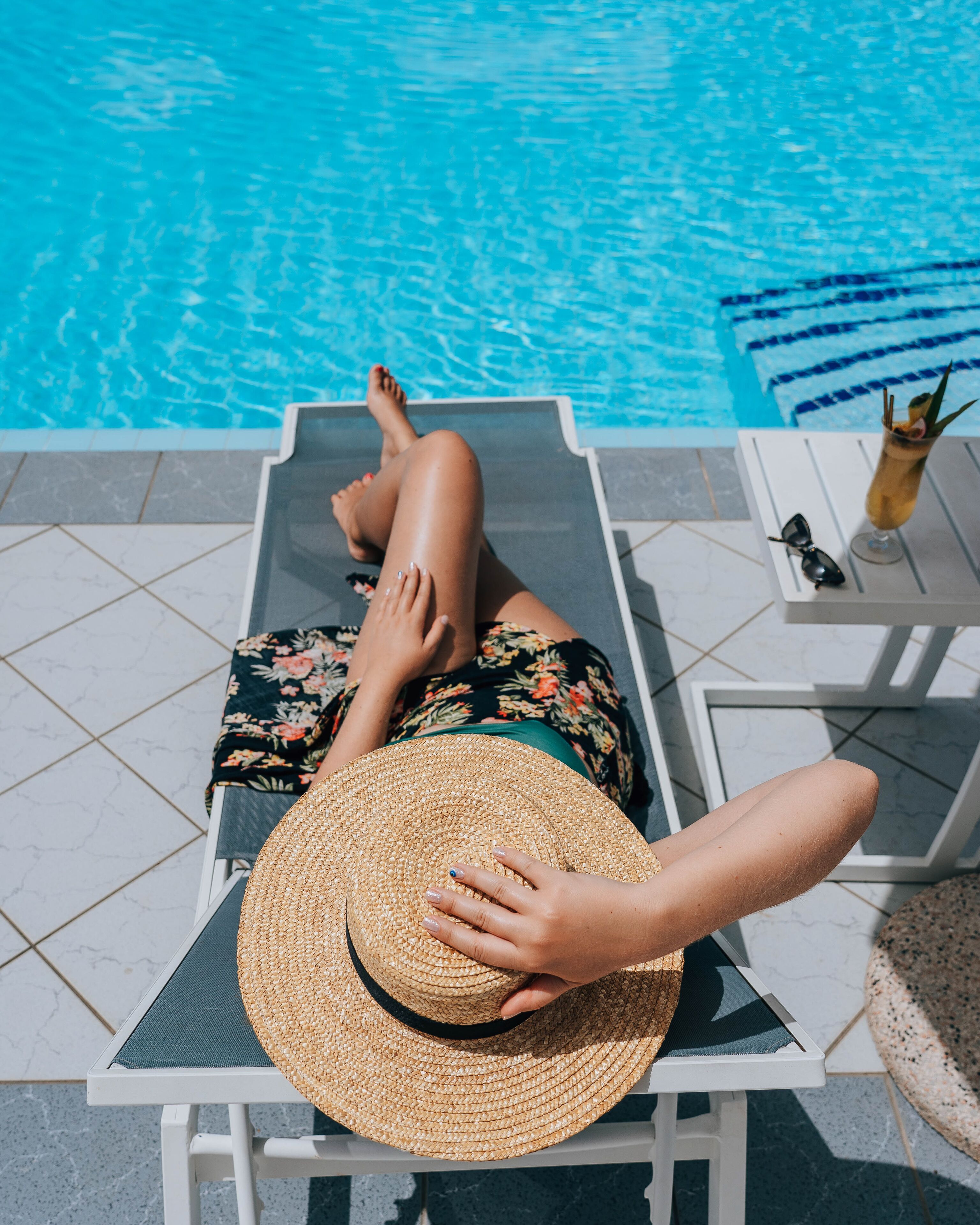 A person lying on a lounge chair by a pool, with a straw hat on their head.
