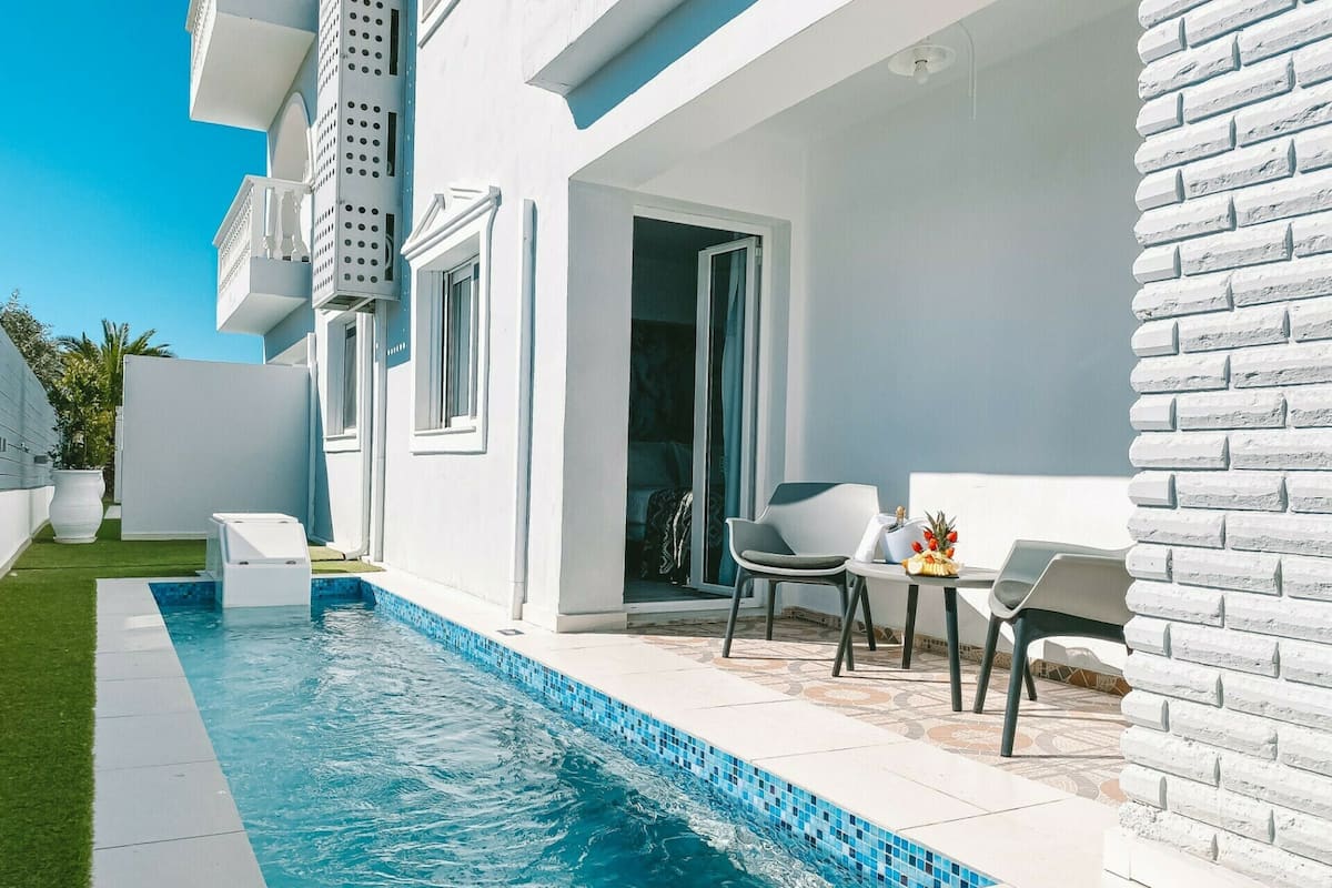 Un área de piscina moderna al aire libre con una piscina azul cristalina, una pequeña zona de estar con una mesa y sillas, y un edificio blanco con balcones.