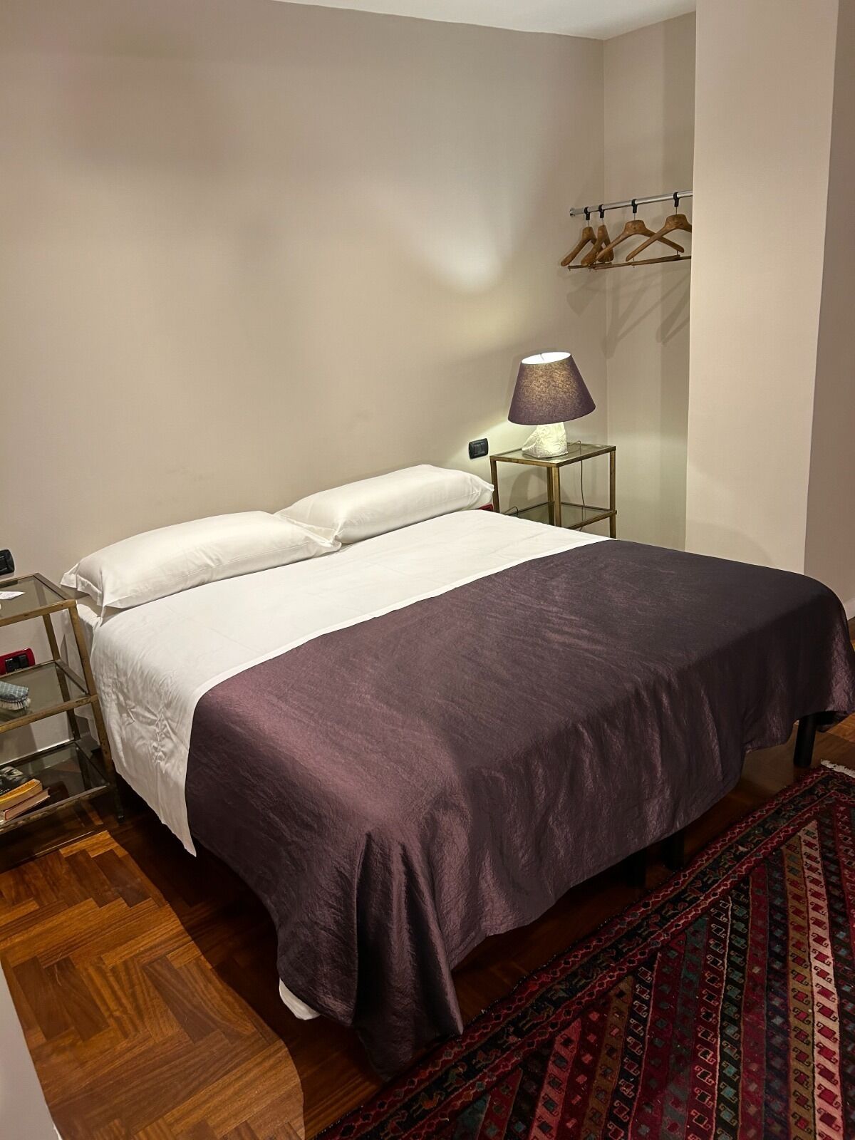 Un letto rifatto con lenzuola bianche e viola, un comodino con una lampada e un attaccapanni a parete.