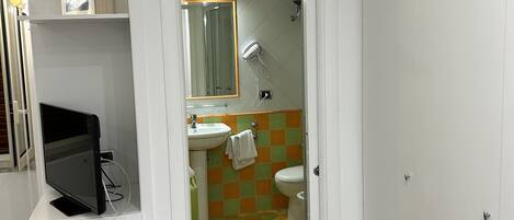 Une salle de bain avec des toilettes, un lavabo et un miroir. On y trouve une étagère avec des objets décoratifs et un livre. Le sol est recouvert d'un motif à carreaux.