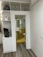 Casa de banho com sanita, lavatório e espelho. Há uma prateleira com objetos decorativos e um livro. O chão tem um padrão riscado.