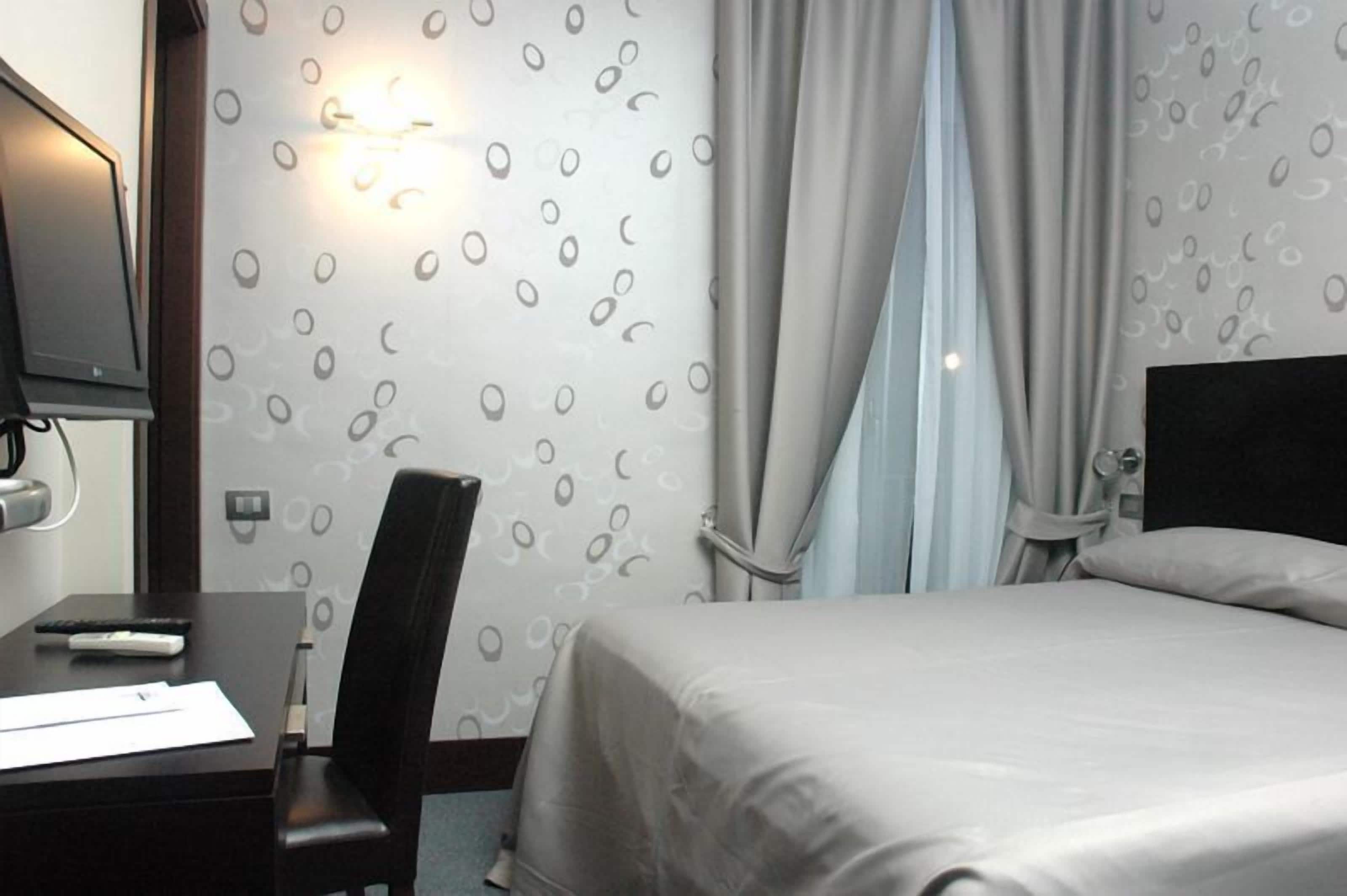 Una habitación de hotel con una cama, un escritorio con silla, un televisor y una lámpara fijada a la pared.