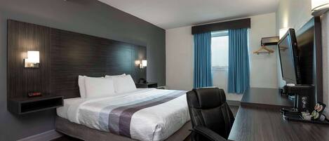 Quarto de hotel com uma cama grande, uma mesa com computador, uma cadeira, televisão e janela com cortinas.