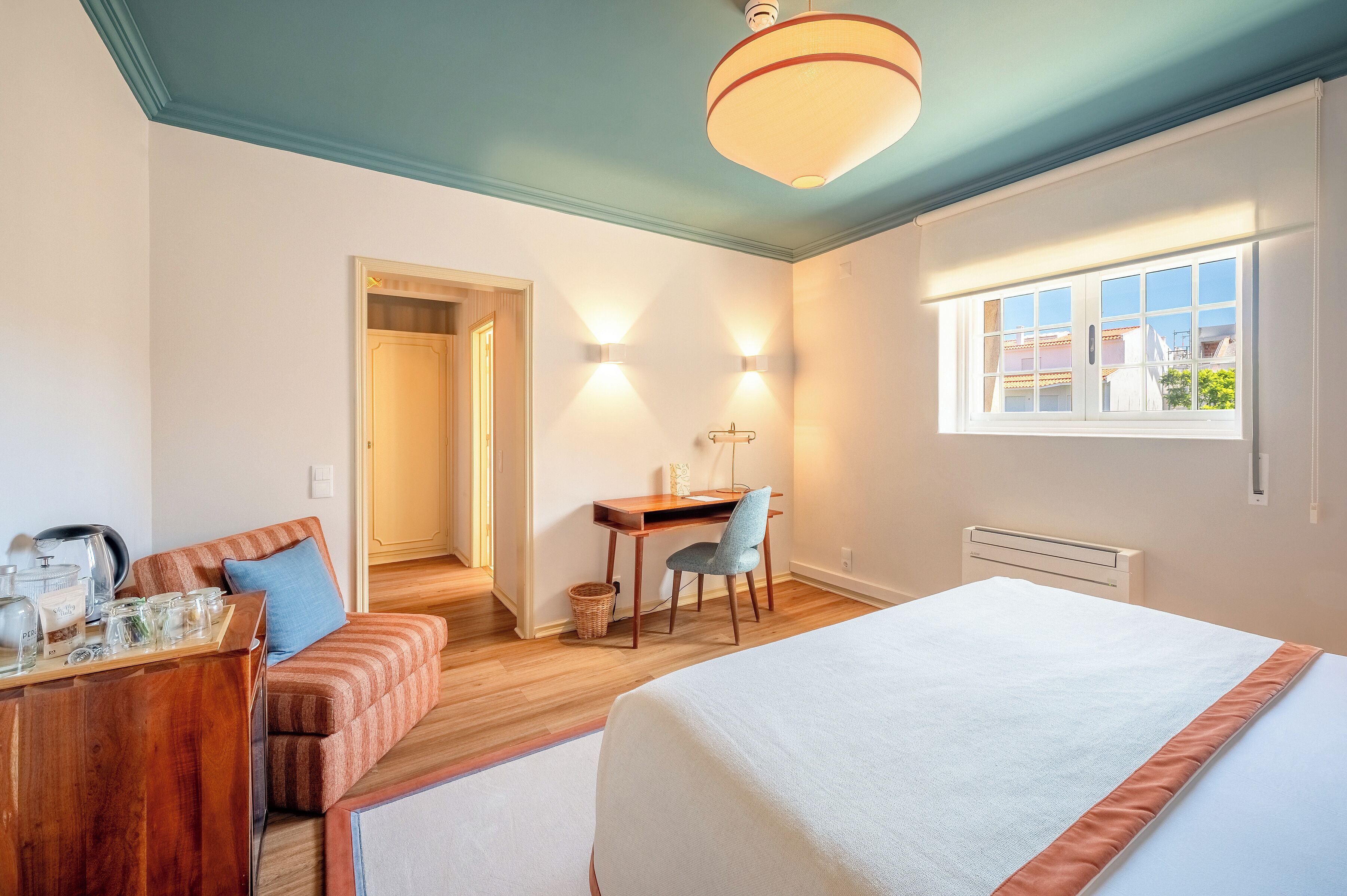 Una habitación de hotel con cama, escritorio, silla y una mesita con objetos encima.