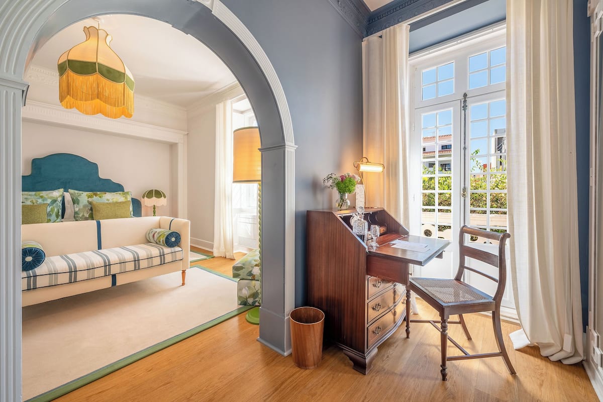 A room with a sofa, a desk with a chair, a lamp, and a view through a window.