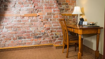 A wooden dining table with a chair, a lamp, and a coffee maker on a carpeted floor with a brick wall in the background.