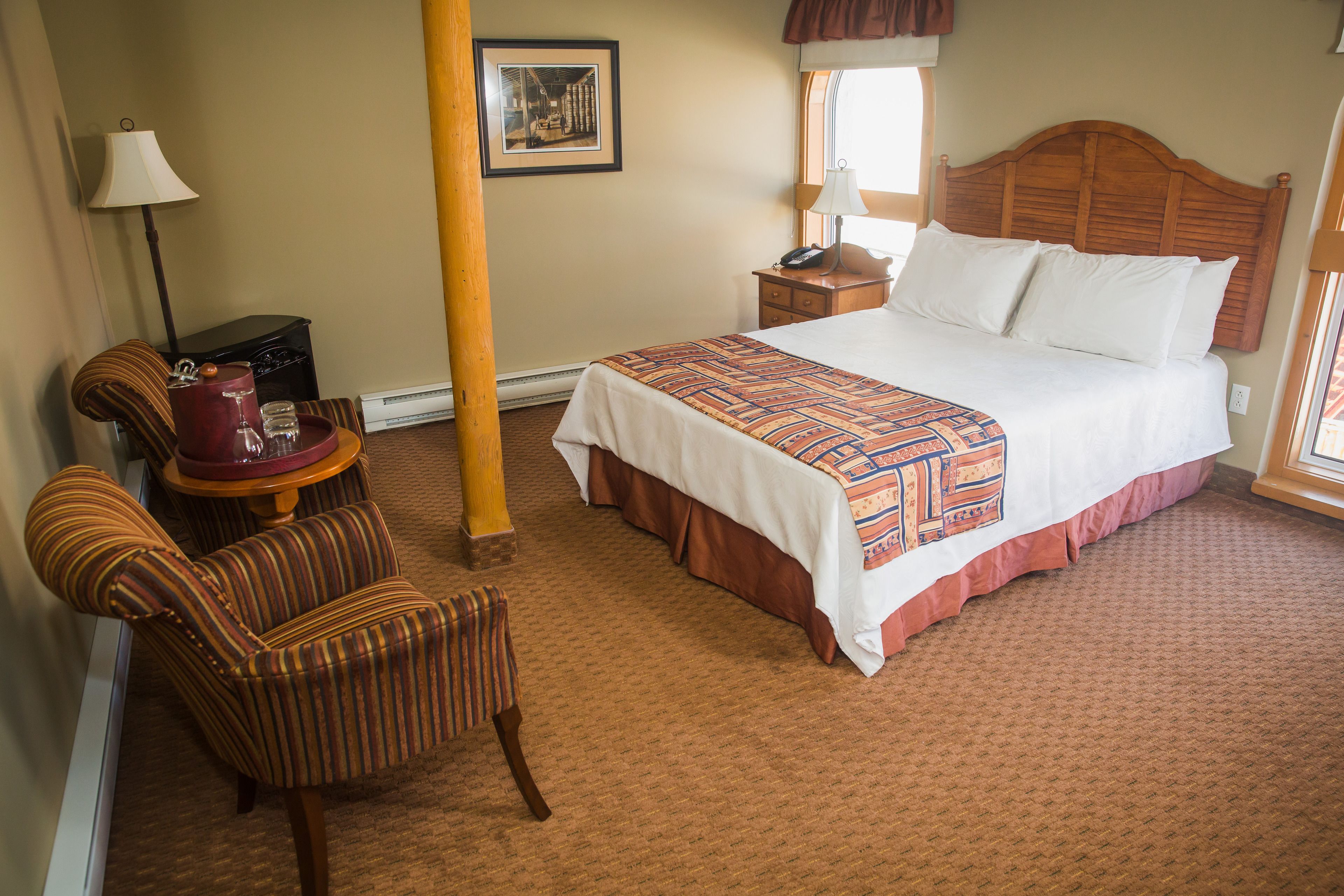 Une chambre d’hôtel avec un grand lit, une table de chevet en bois, une lampe, une chaise et une petite table sur laquelle se trouve un pichet rouge.
