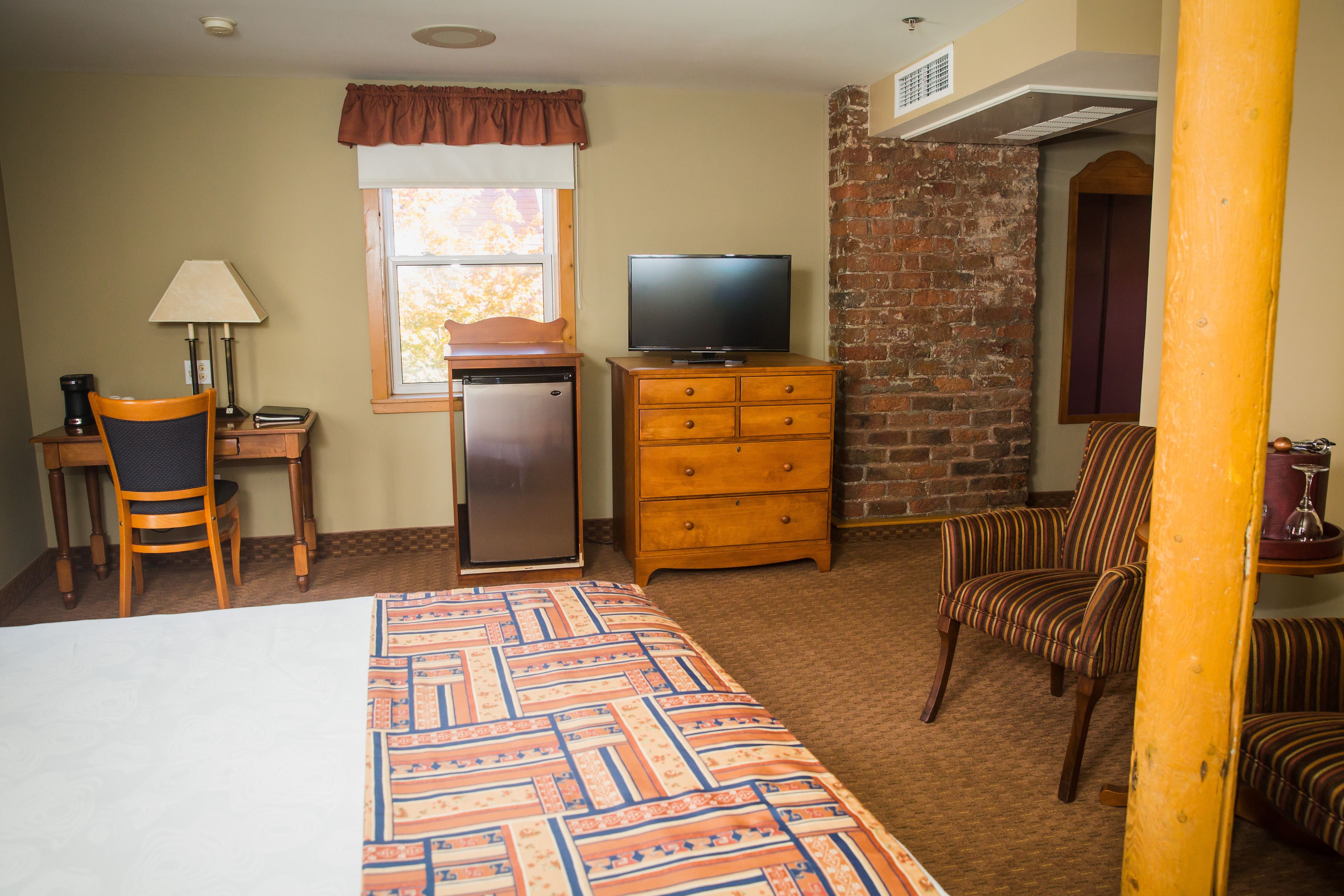 A hotel room with a bed, a desk with a lamp, a television, a refrigerator, and a brick wall.