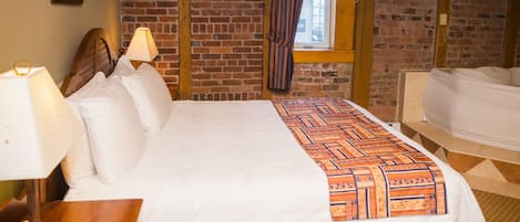 A hotel room with a large bed, a wooden nightstand with a lamp, a brick wall, and a window with curtains.