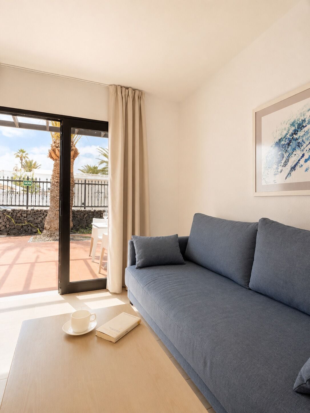Una habitación moderna con un sofá gris, una mesa de centro blanca, un cuadro abstracto enmarcado y vistas a palmeras a través de una puerta de cristal.