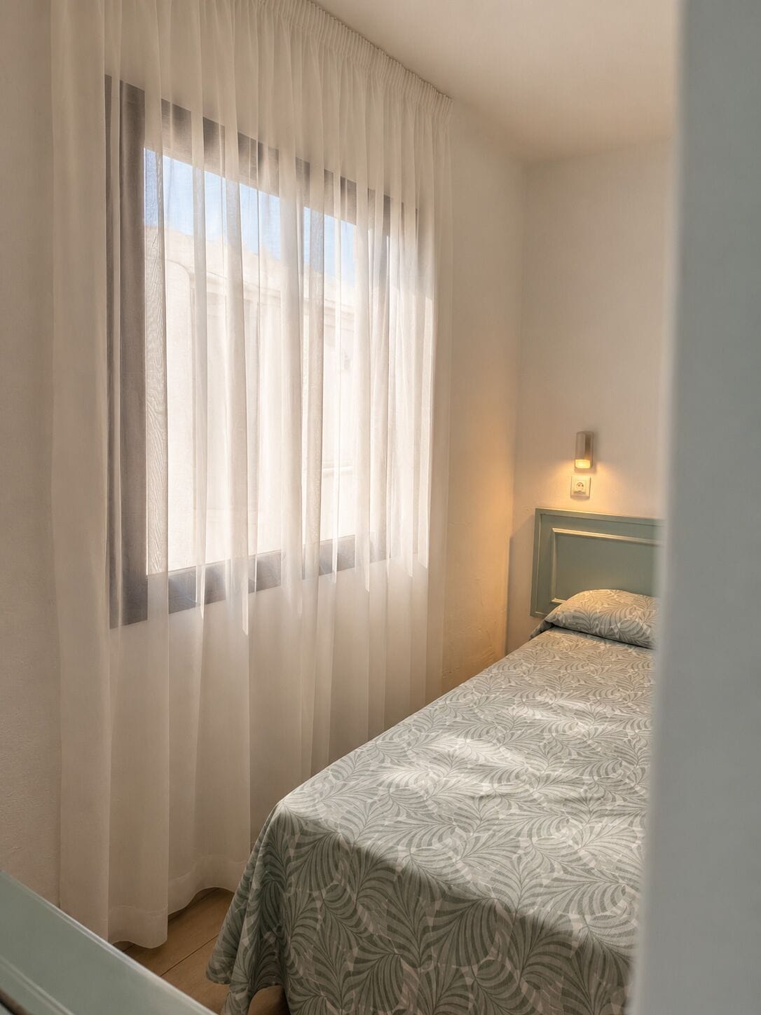 Et soveværelse med en seng, et vindue med lette gardiner og en lampe ophængt på væggen.