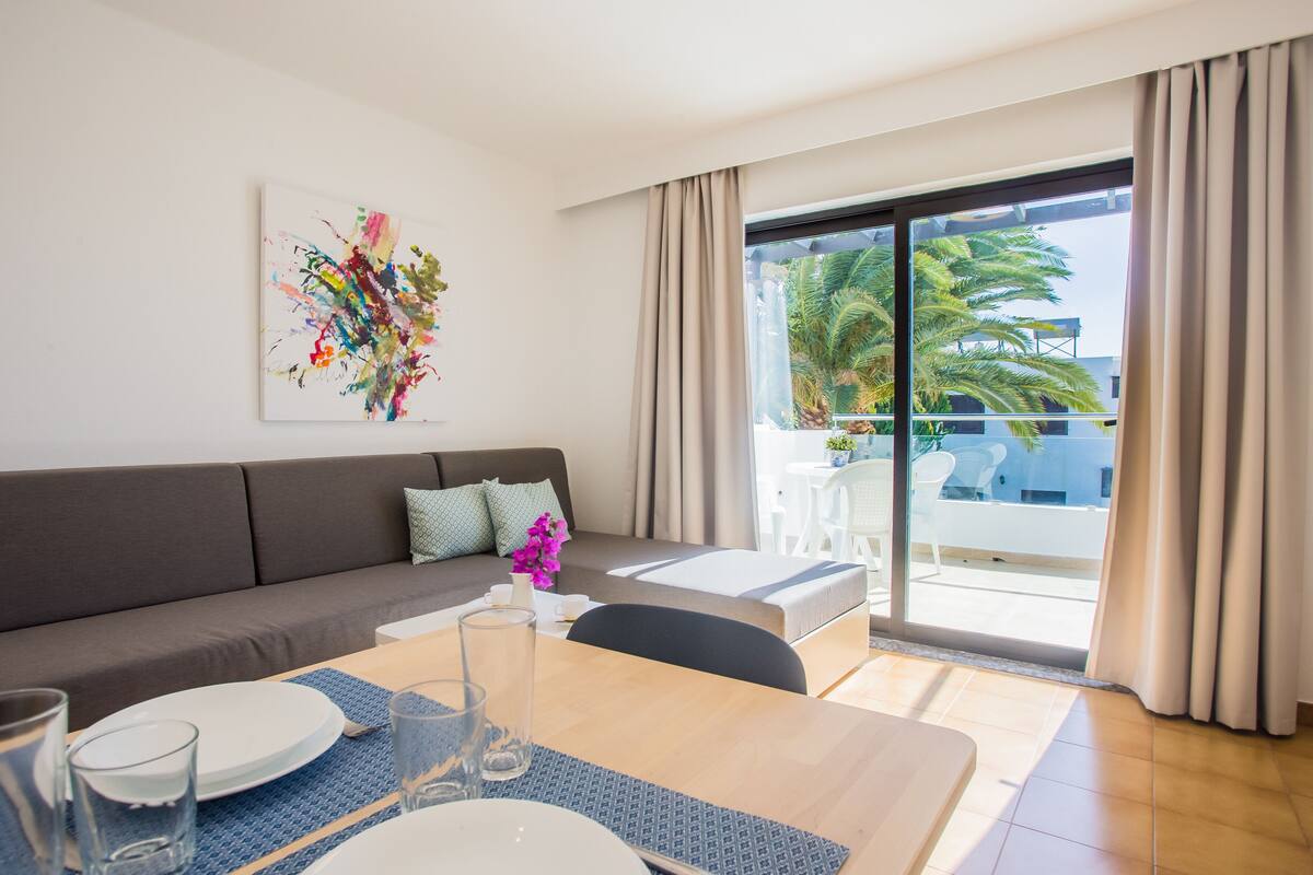 Un salon moderne avec un canapé, une table à manger et un balcon offrant une vue sur les palmiers.