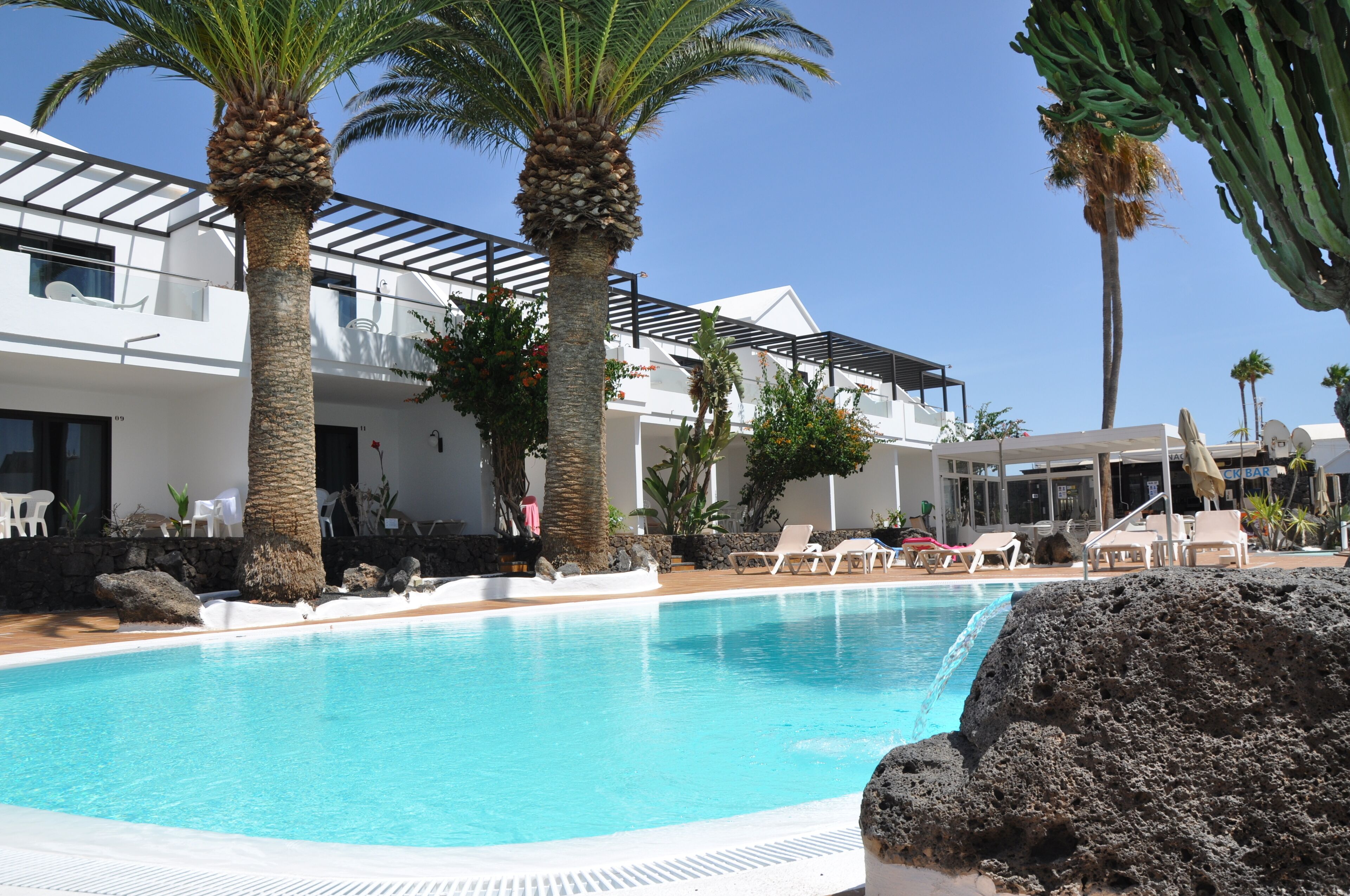 A pool area with lounge chairs, palm trees, and a building with a balcony.