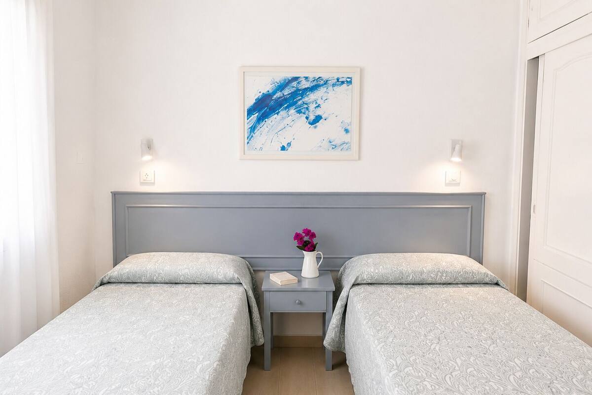 Une chambre double avec deux lits, une table de chevet ornée d’un vase de fleurs, un tableau abstrait encadré et des lampes fixées au mur.