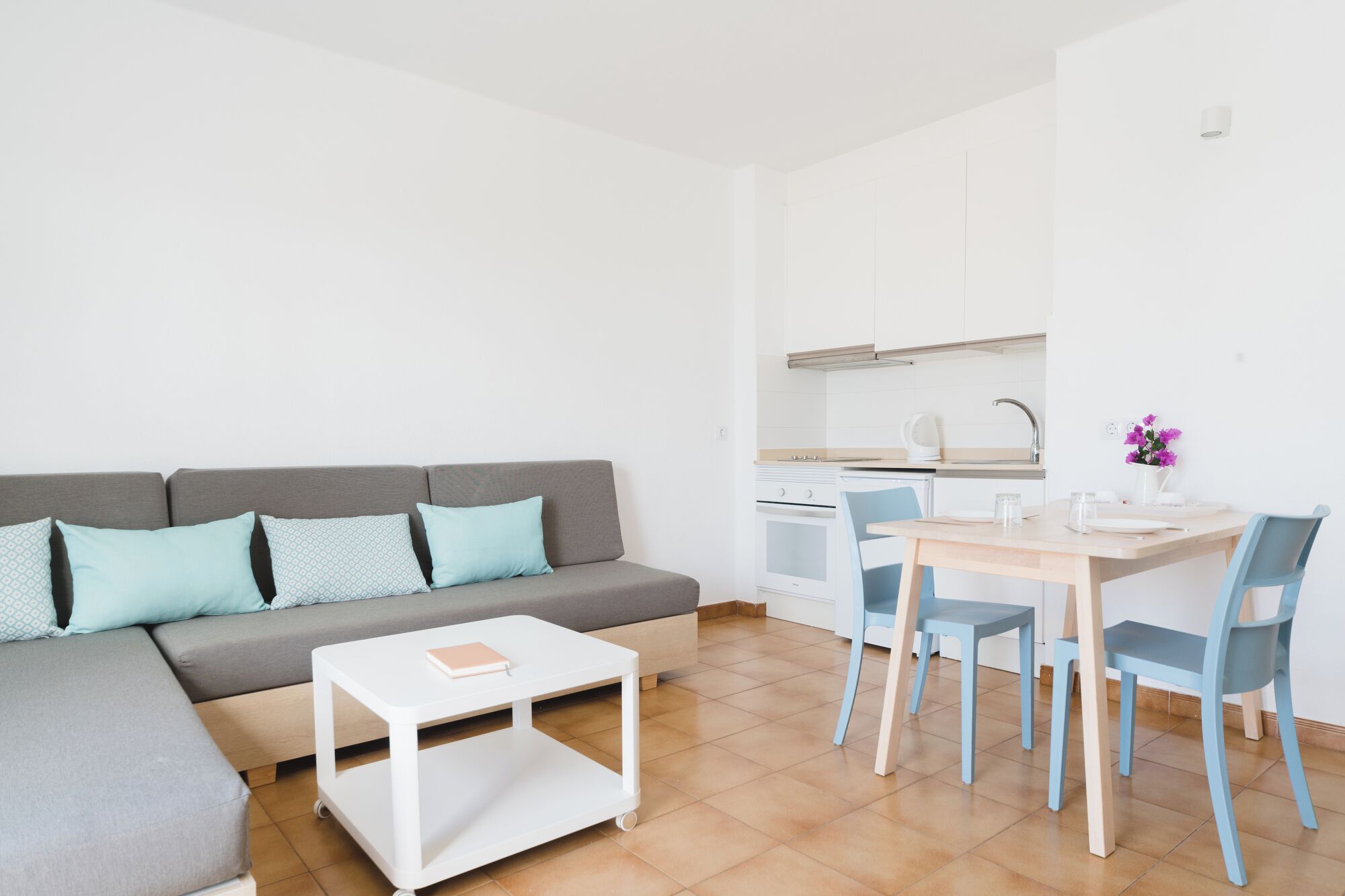 A modern, compact living space with a grey sofa, a small white table, and a dining area with a wooden table and blue chairs.