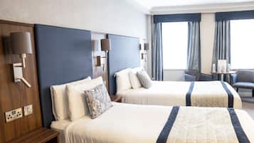 Een hotelkamer met twee bedden, een houten nachtkastje, een kleine tafel met stoel en een groot raam met gordijnen.