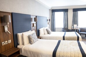 A hotel room with two beds, a wooden nightstand, a small table with a chair, and a large window with curtains.