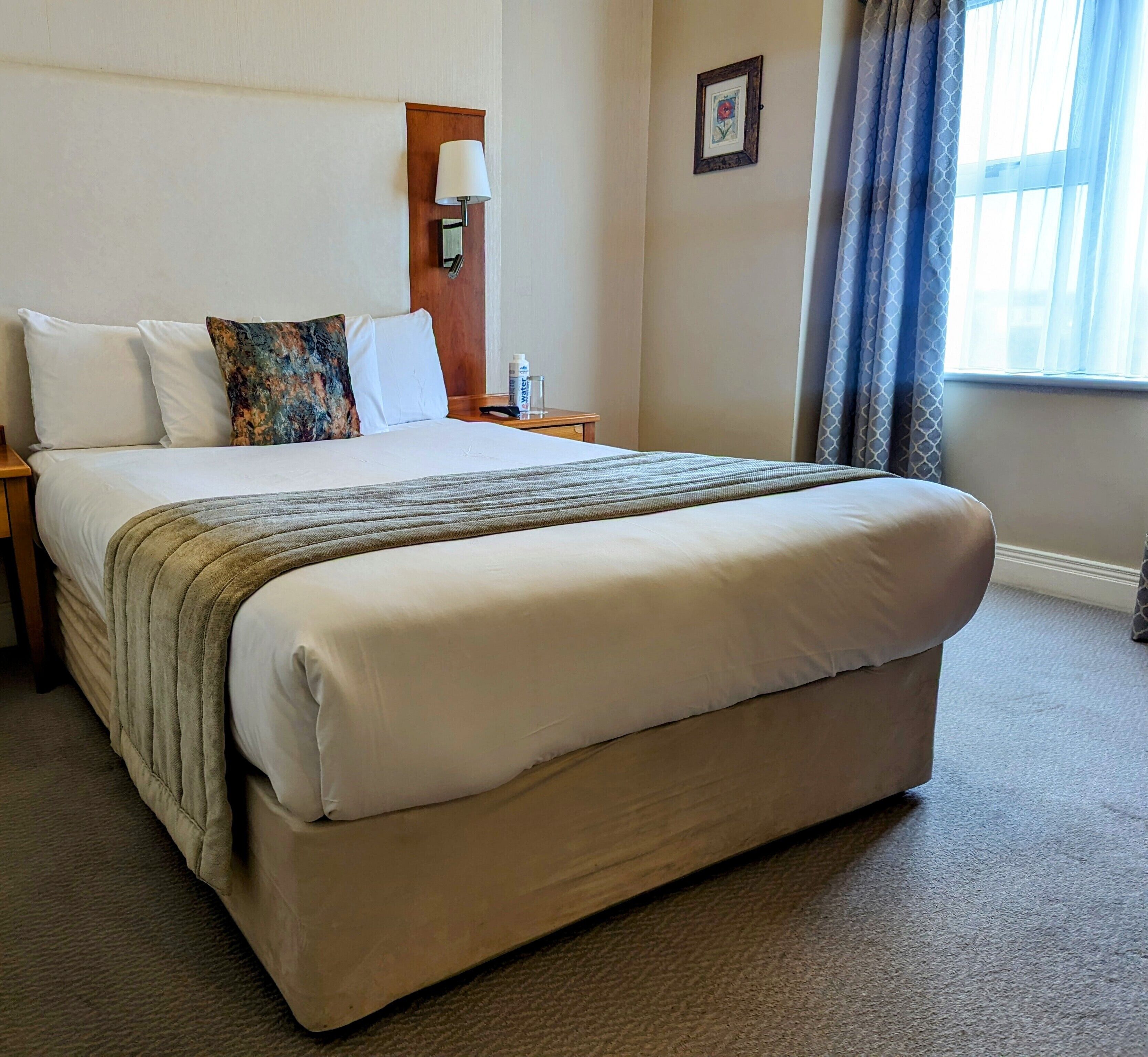 A neatly made bed with a beige comforter and pillows, a bedside table with a lamp and a bottle of water, and a window with curtains.
