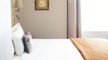 Una cama bien hecha con cubrecama blanco y una manta naranja texturizada. Encima de la cama hay dos cuadros enmarcados, y a la derecha, una ventana con cortina estampada.