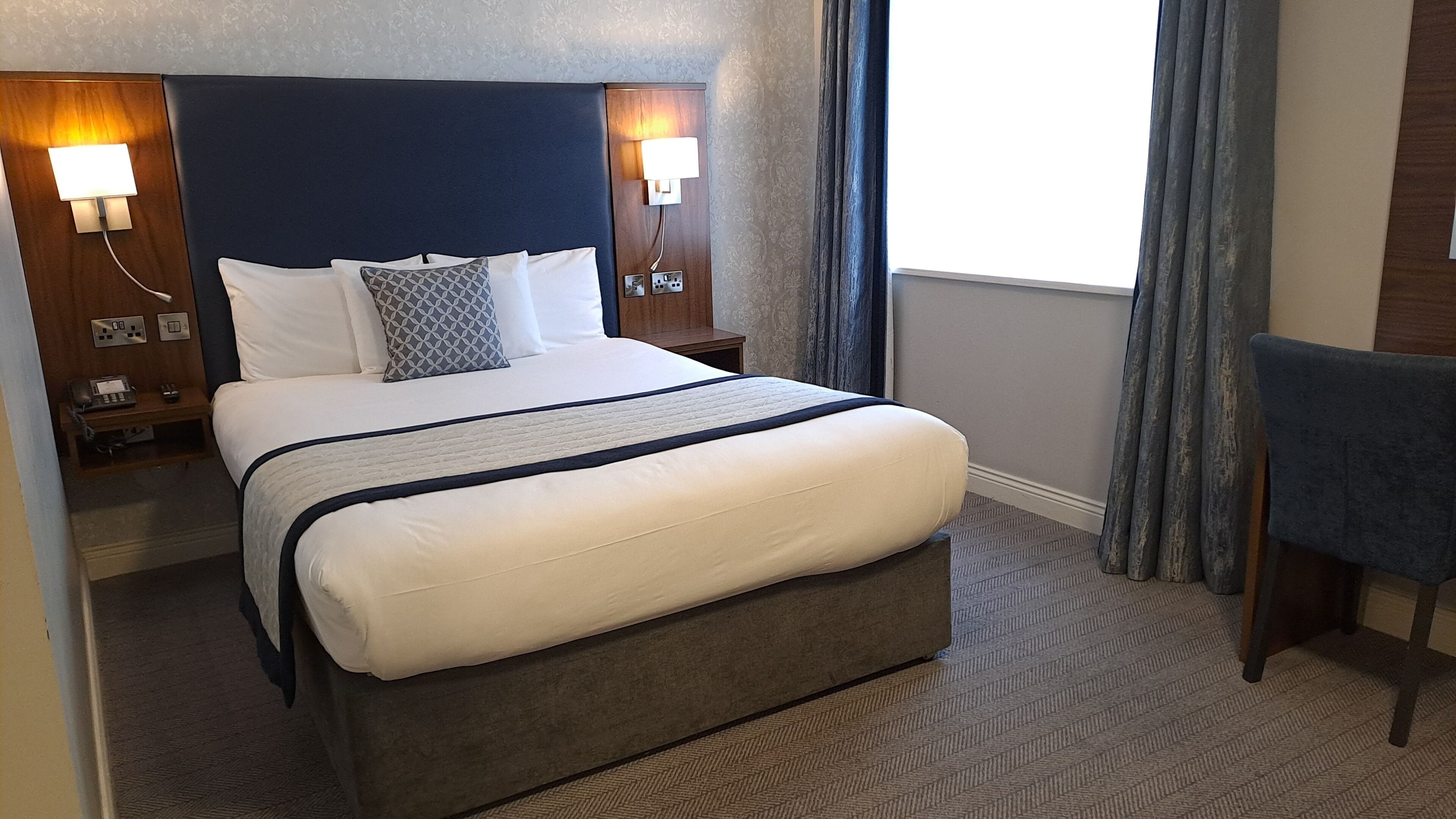Una habitación de hotel con una cama grande, dos mesitas de noche con lámparas, una silla y vistas al exterior.
