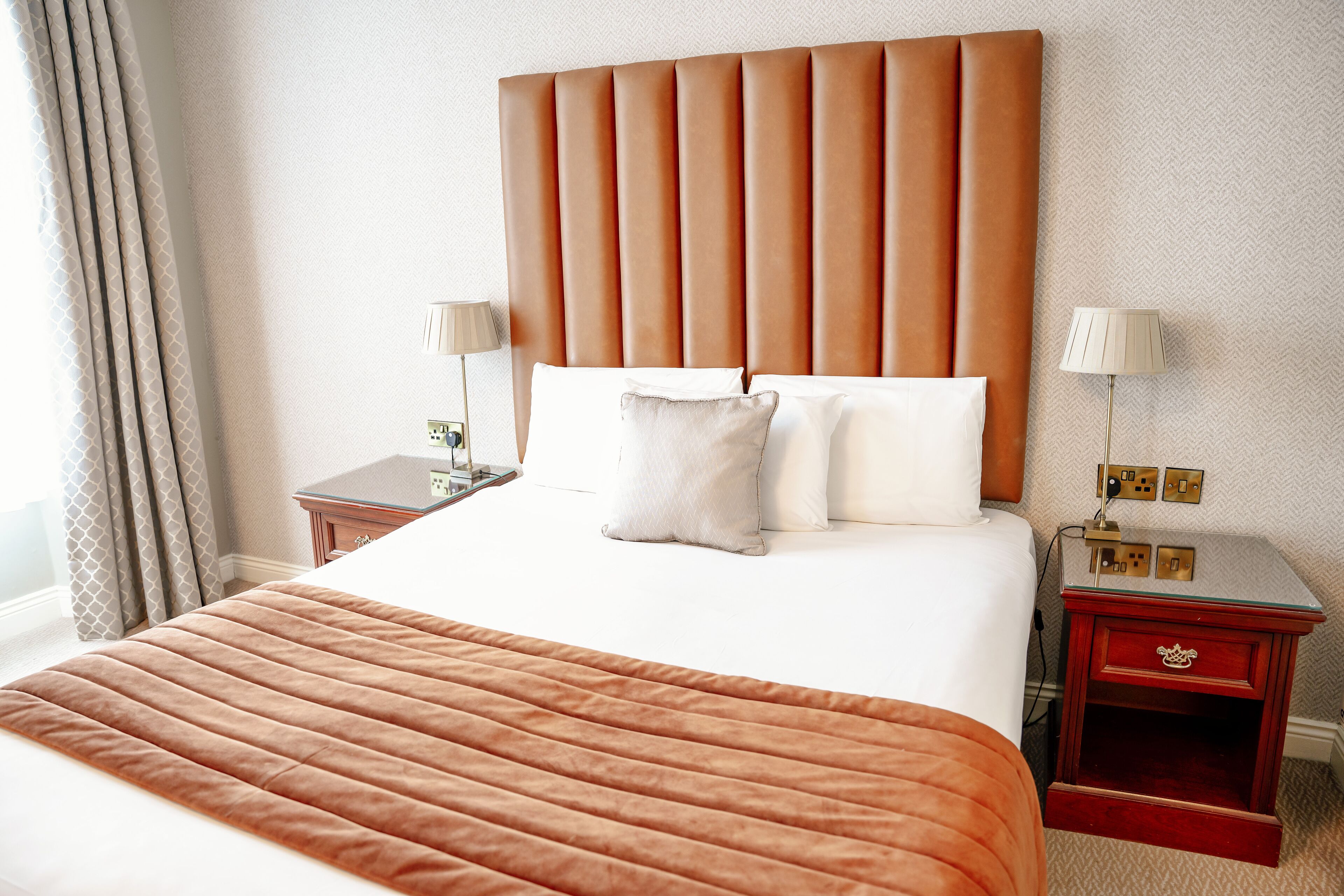 Quarto de hotel com uma cama grande, cabeceira, duas mesas de cabeceira e uma mesinha de apoio.