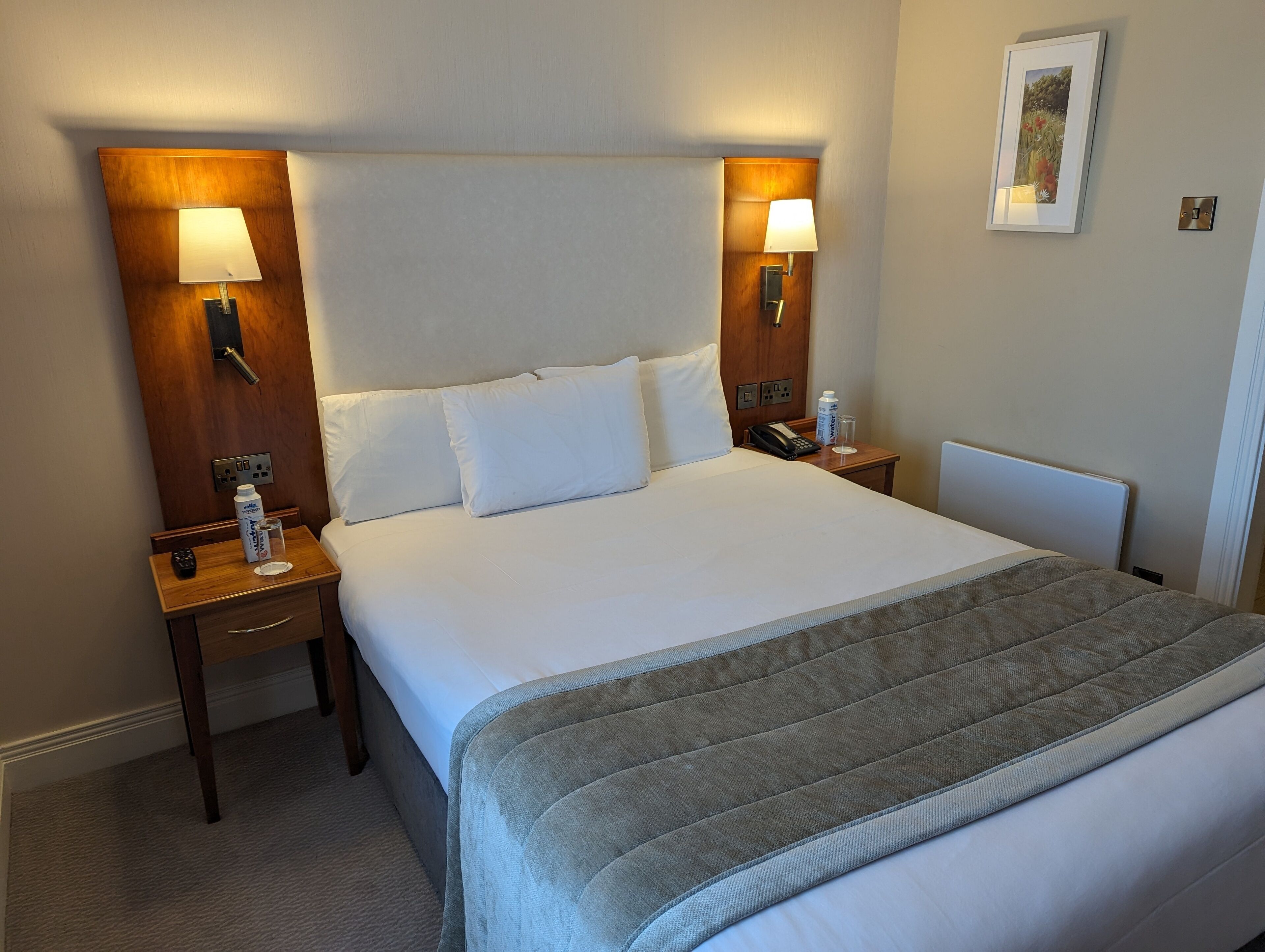A hotel room with a large bed, two bedside tables, a nightstand, a bedside lamp, a framed picture on the wall, and a thermostat.
