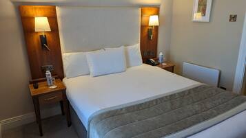 A hotel room with a large bed, two bedside tables, a nightstand, a bedside lamp, a framed picture on the wall, and a thermostat.