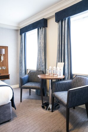 A hotel room with a bed, two armchairs, a small table with water bottles, a lamp, and a view through large windows with curtains. - The Western Hotel (Galway)