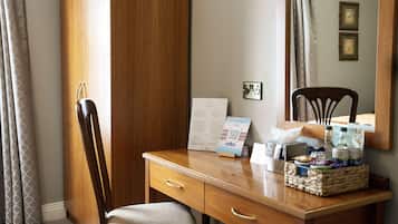 A hotel room with a wooden desk, a chair, a mirror, a framed picture, a sign, and a basket of toiletries.