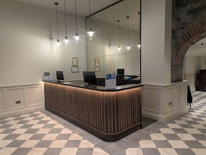 A reception desk with a check-in counter, overhead pendant lights, framed certificates on the wall, and a check-in station with a computer monitor. - The Western Hotel (Galway)