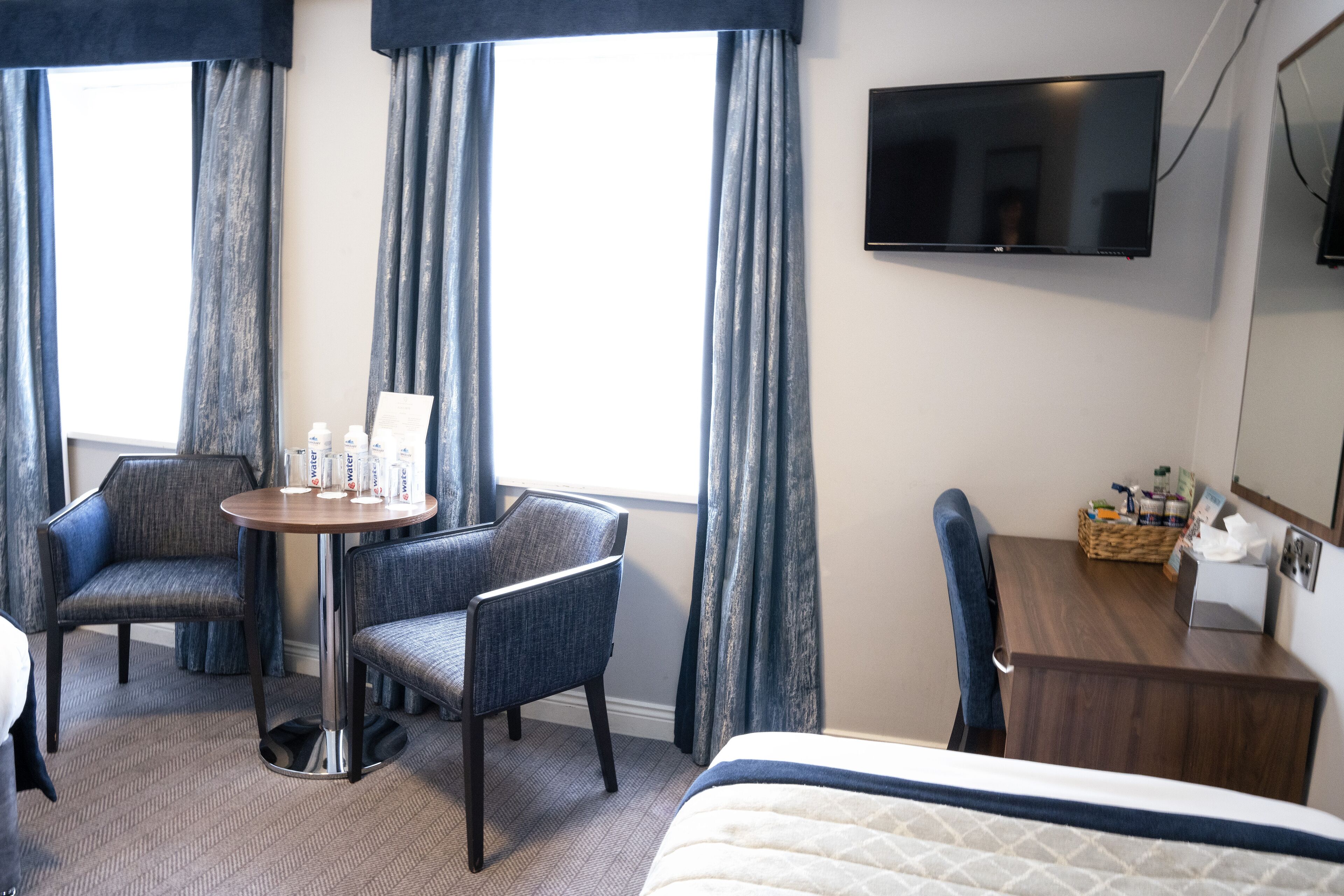 Une chambre d’hôtel avec un lit, deux fauteuils, une petite table ronde avec des bouteilles d’eau, un téléviseur fixé au mur et un bureau en bois avec un panier et des mouchoirs.
