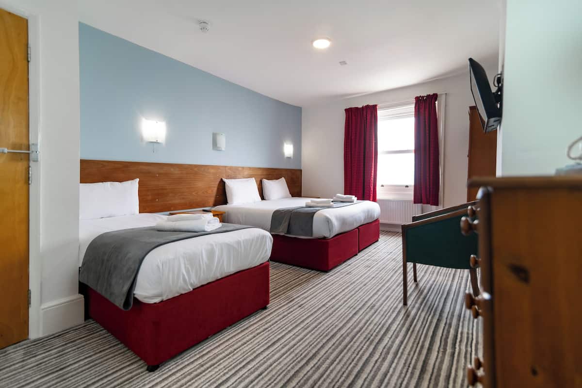 A hotel room with two beds, a wooden headboard, a television, and a window with red curtains.