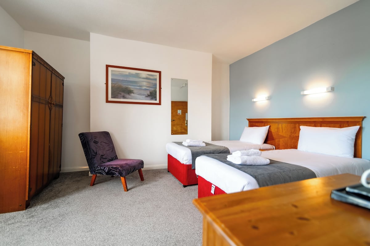 A hotel room with two beds, a wooden wardrobe, a chair, a small table, and a painting on the wall.