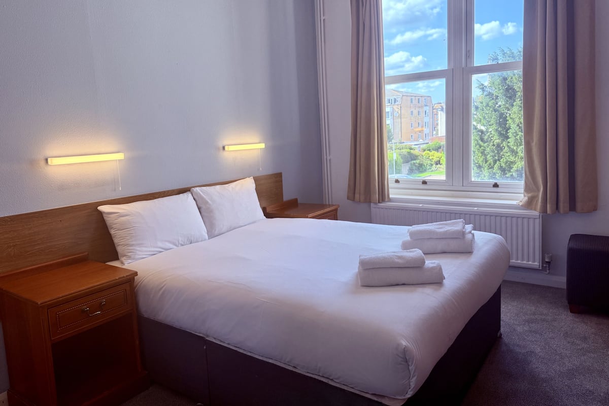 A hotel room with a large bed, two bedside tables, a window with curtains, and a view of buildings and trees.