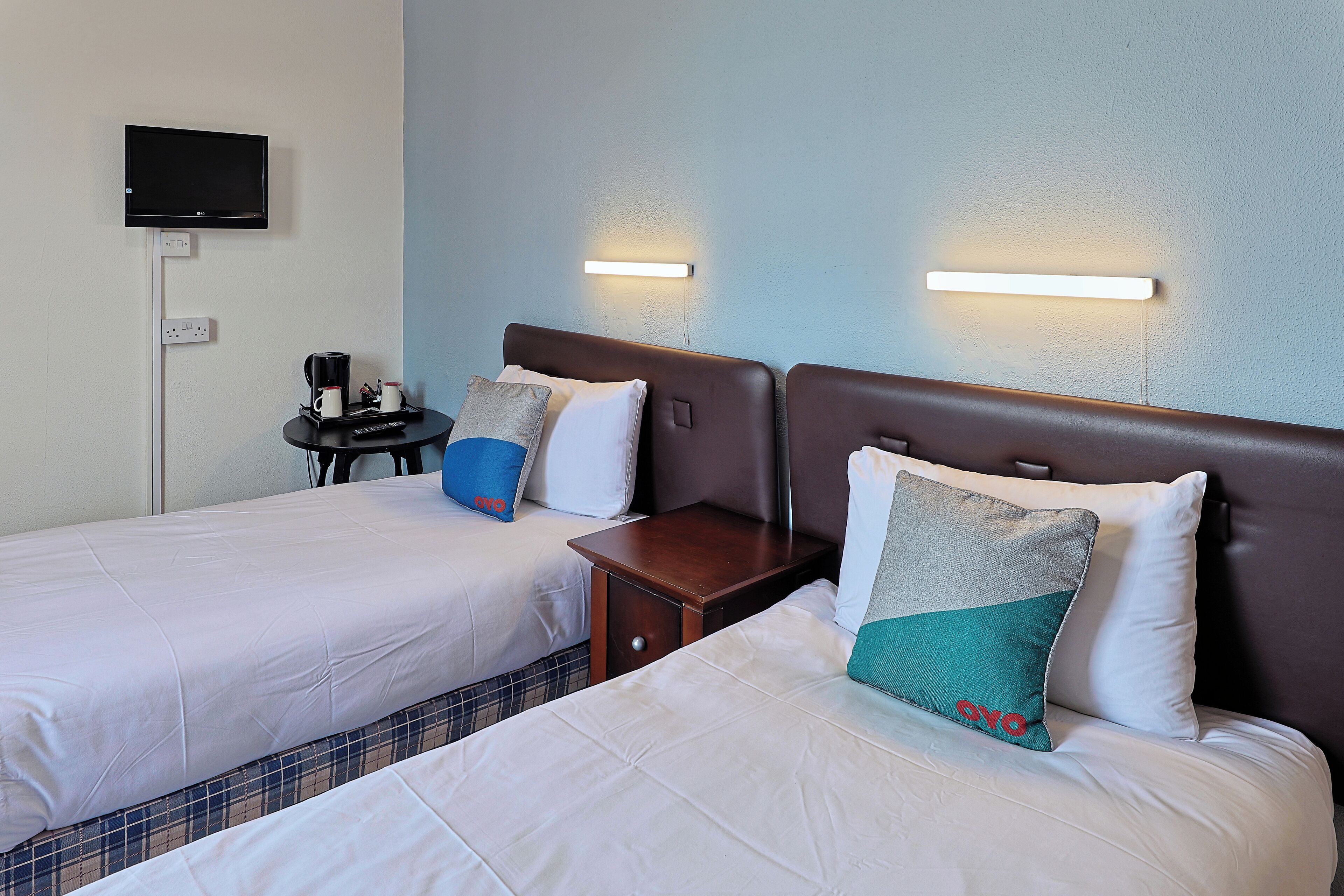 Ein Hotelzimmer mit zwei Betten, einem an der Wand montierten Fernseher, Nachttischen und einem kleinen Tisch mit einer Kaffeemaschine.