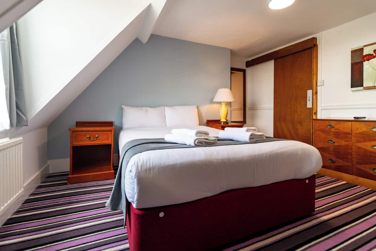A hotel room with a bed, a nightstand, a lamp, a wooden dresser, and a window with curtains.