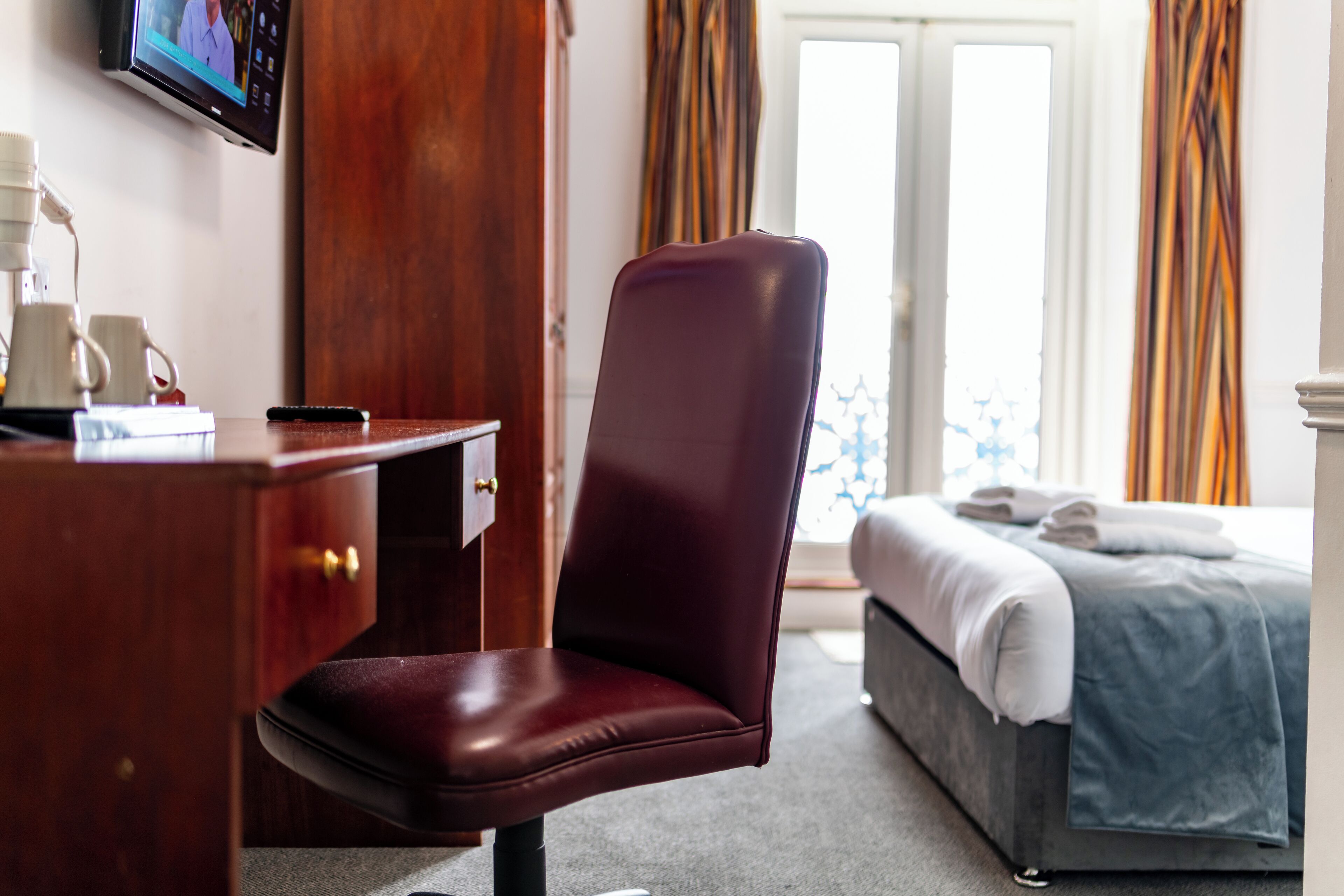 A hotel room with a desk, a chair, a bed, and a TV on the wall.