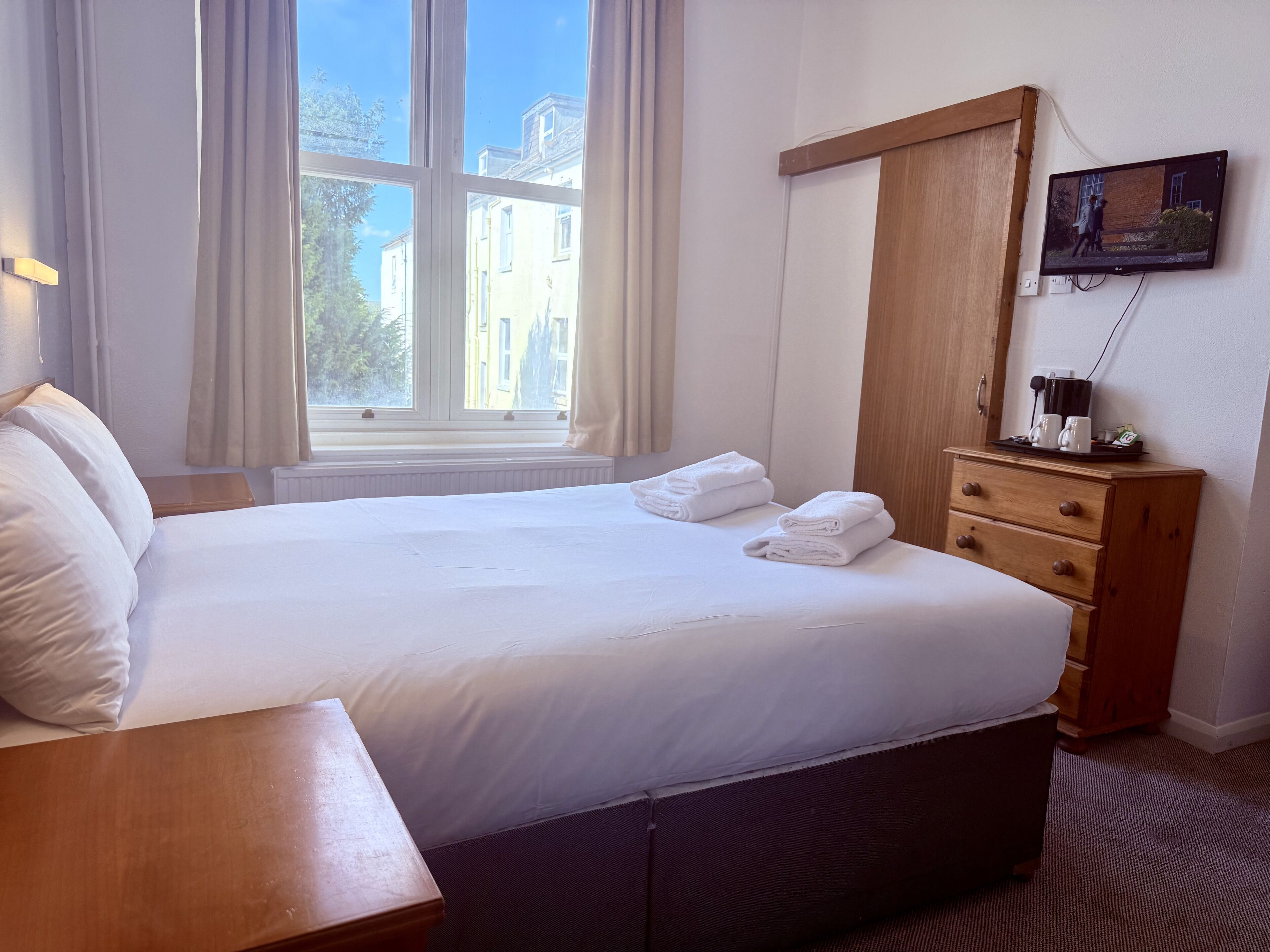 Ein Hotelzimmer mit einem Bett, einem Nachttisch aus Holz, einer Kommode, einem an der Wand montierten Fernseher und einem Fenster mit Vorhängen.