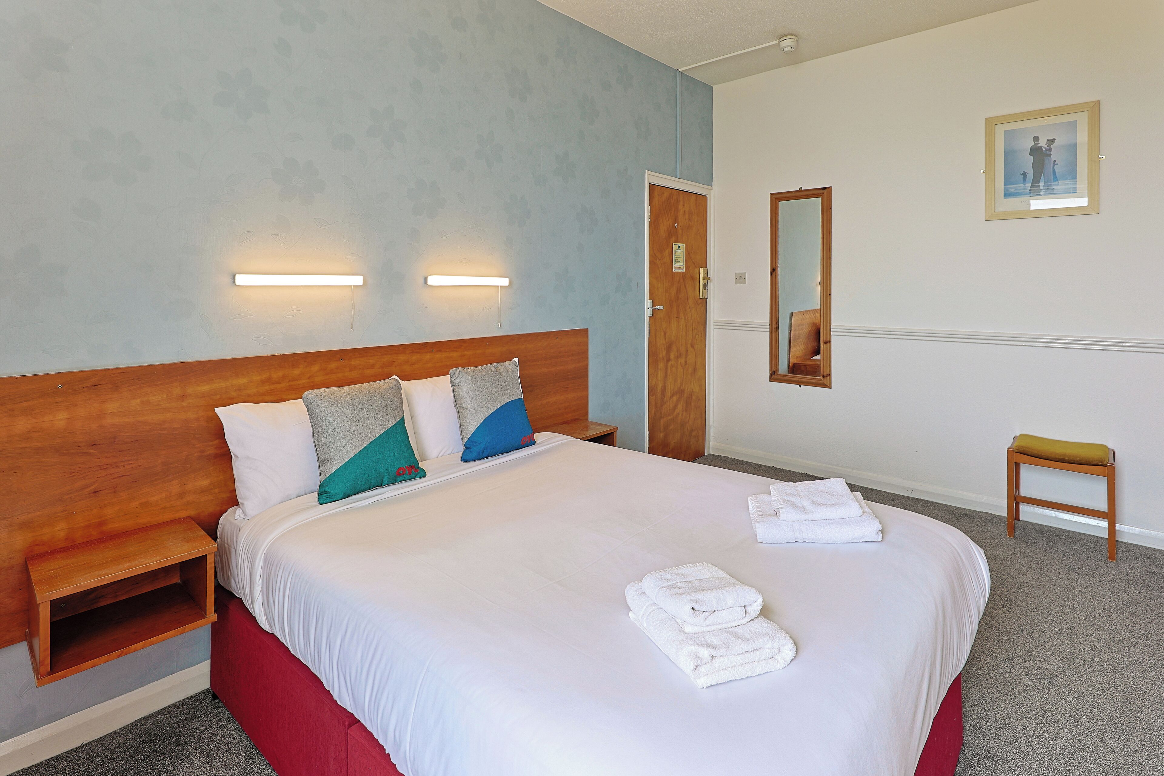 Quarto de hotel com uma cama grande, mesas de cabeceira, painel de madeira na cabeceira, um pequeno banquinho, um espelho e um quadro emoldurado na parede.