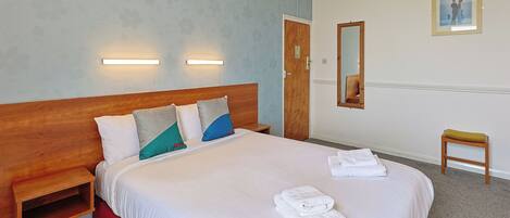 Quarto de hotel com uma cama grande, mesas de cabeceira, painel de madeira na cabeceira, um pequeno banquinho, um espelho e um quadro emoldurado na parede.