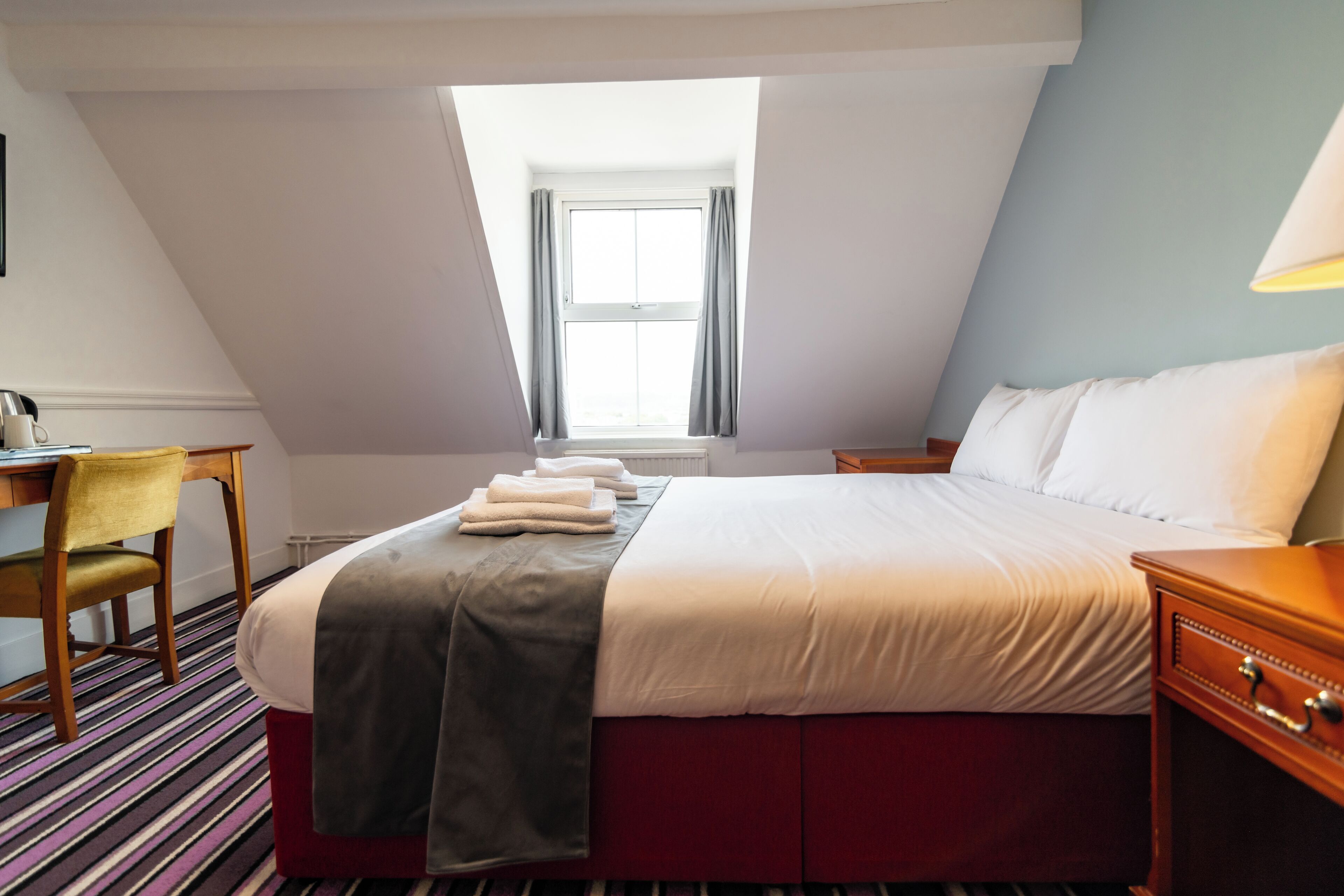 A hotel room with a bed, a desk, a chair, a window, and a lamp.