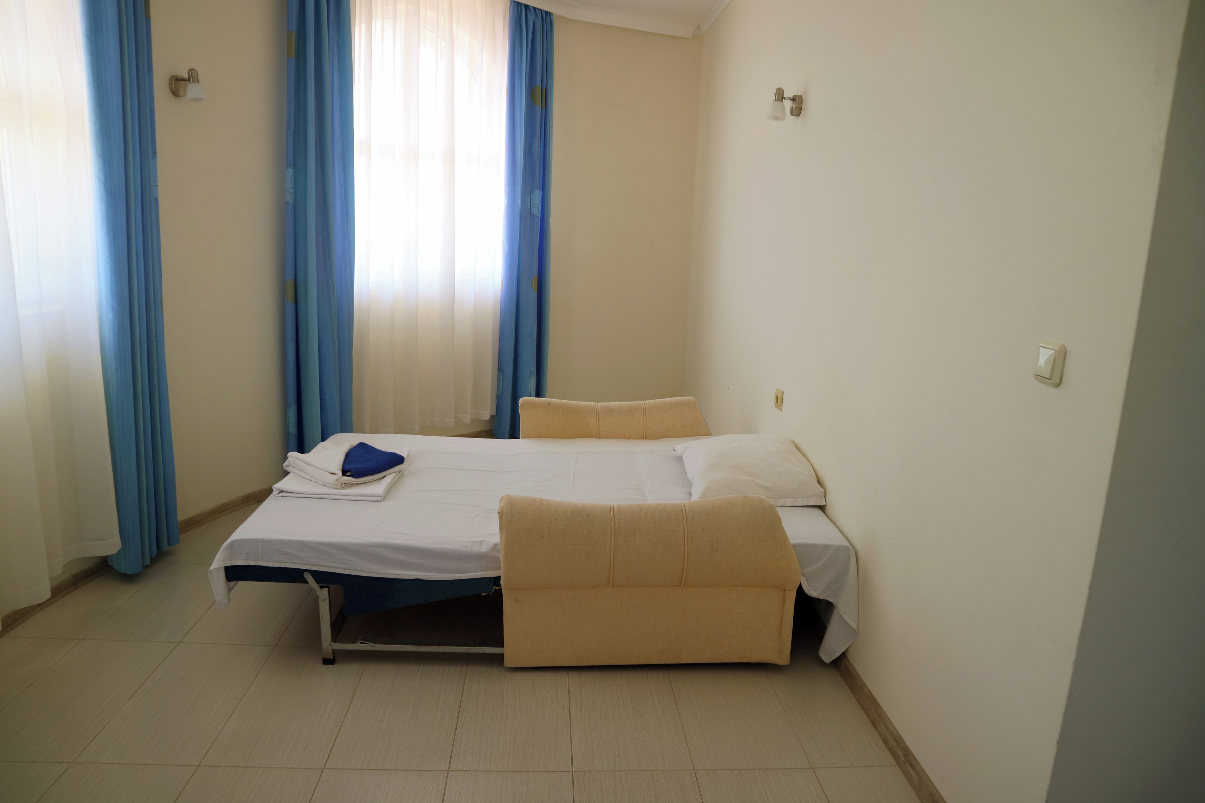 A single-bed room with a pull-out sofa, a bedside table, and a window with blue curtains.