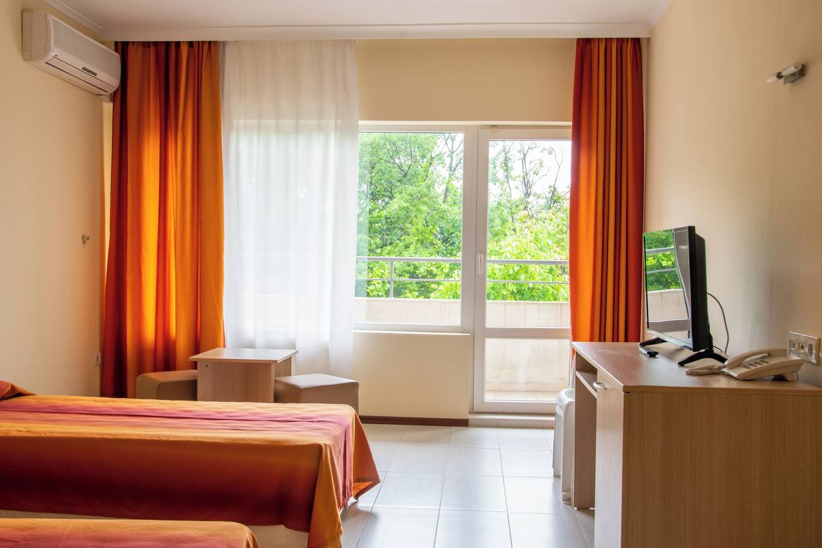 A hotel room with two beds, a desk with a computer and phone, and a balcony with orange curtains.