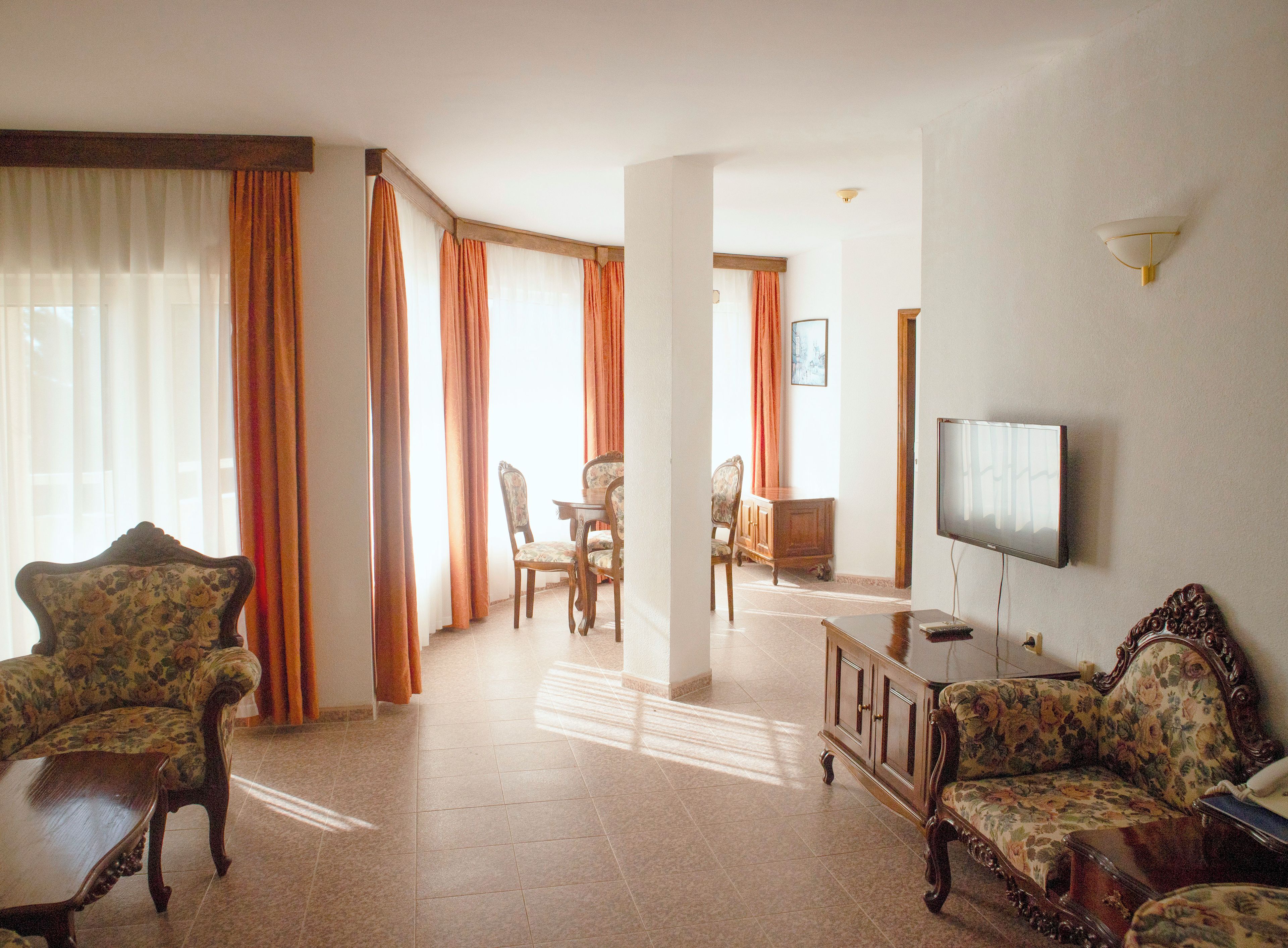 A living room with a television, floral-patterned sofas, a dining area with chairs, and a view through a window with curtains.