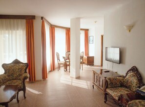 A living room with a television, floral-patterned sofas, a dining area with chairs, and a view through a window with curtains. - Park Hotel Bellevue (Golden Sands)