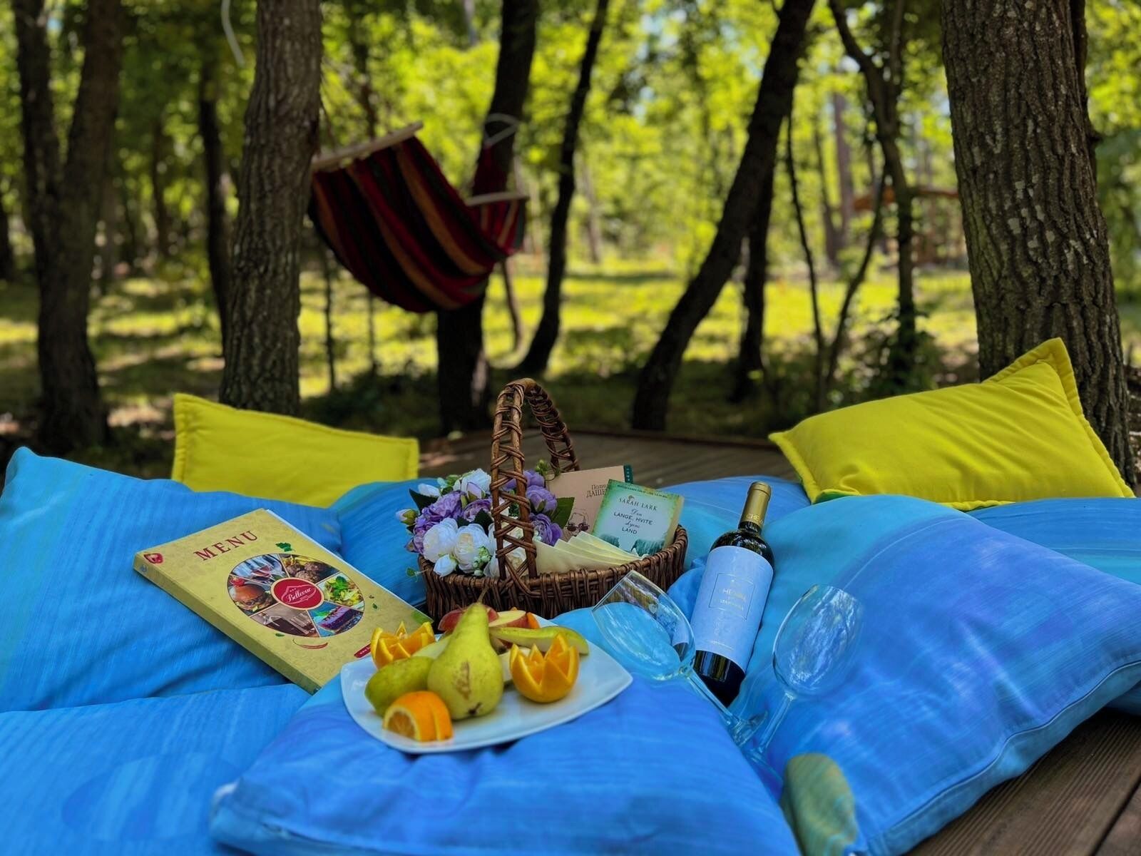 A picnic setup on a blue blanket in a forest with a wicker basket, a menu, a bottle of champagne, and a plate of fruit.
