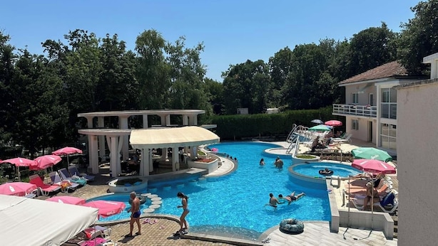 Una piscina con sillas de descanso y sombrillas, rodeada de árboles y un edificio.